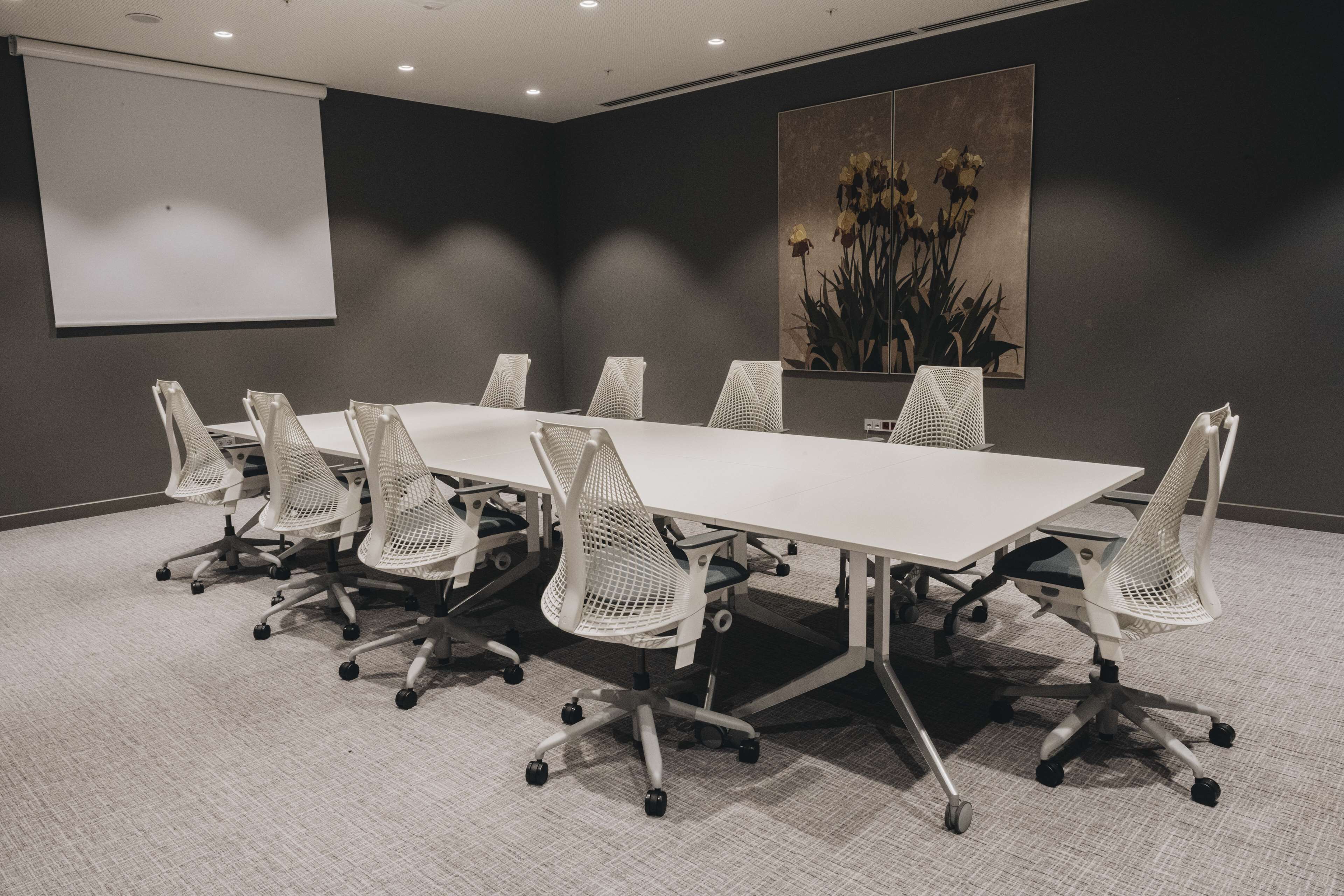 A conference room with a long table, white mesh chairs, a projector screen, and a large floral painting on the wall.