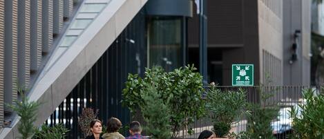 Un café al aire libre con mesas y sillas, plantas en macetas y un edificio moderno al fondo.