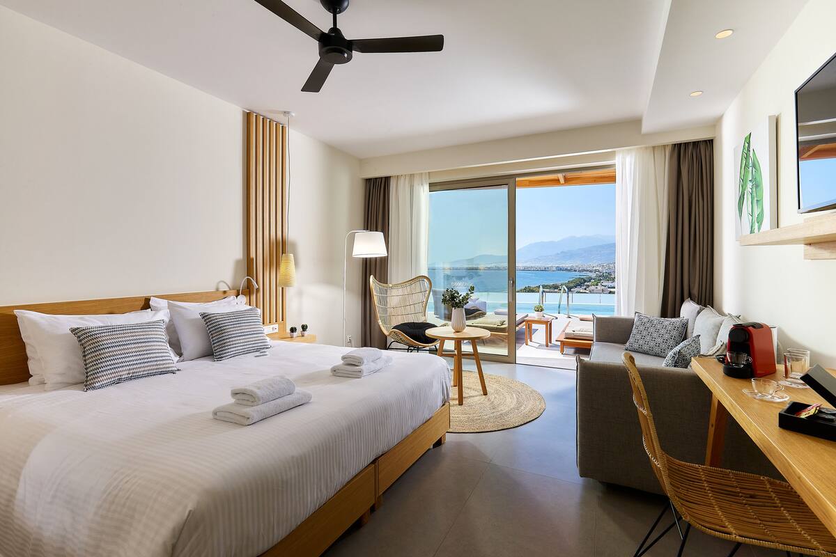 A hotel room with a bed, a desk, a chair, a table, and a view of the sea.
