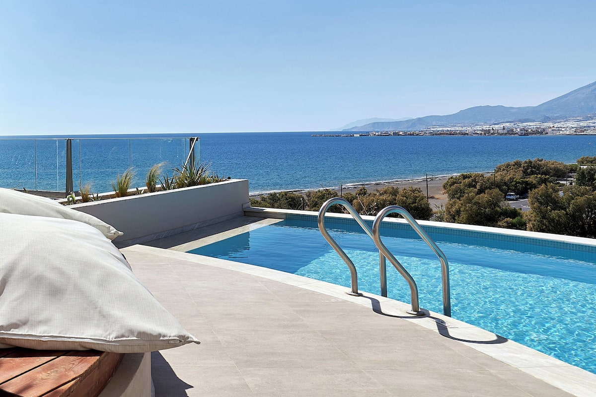 A pool with a view of the sea and mountains, a wooden bench, and a white pillow.