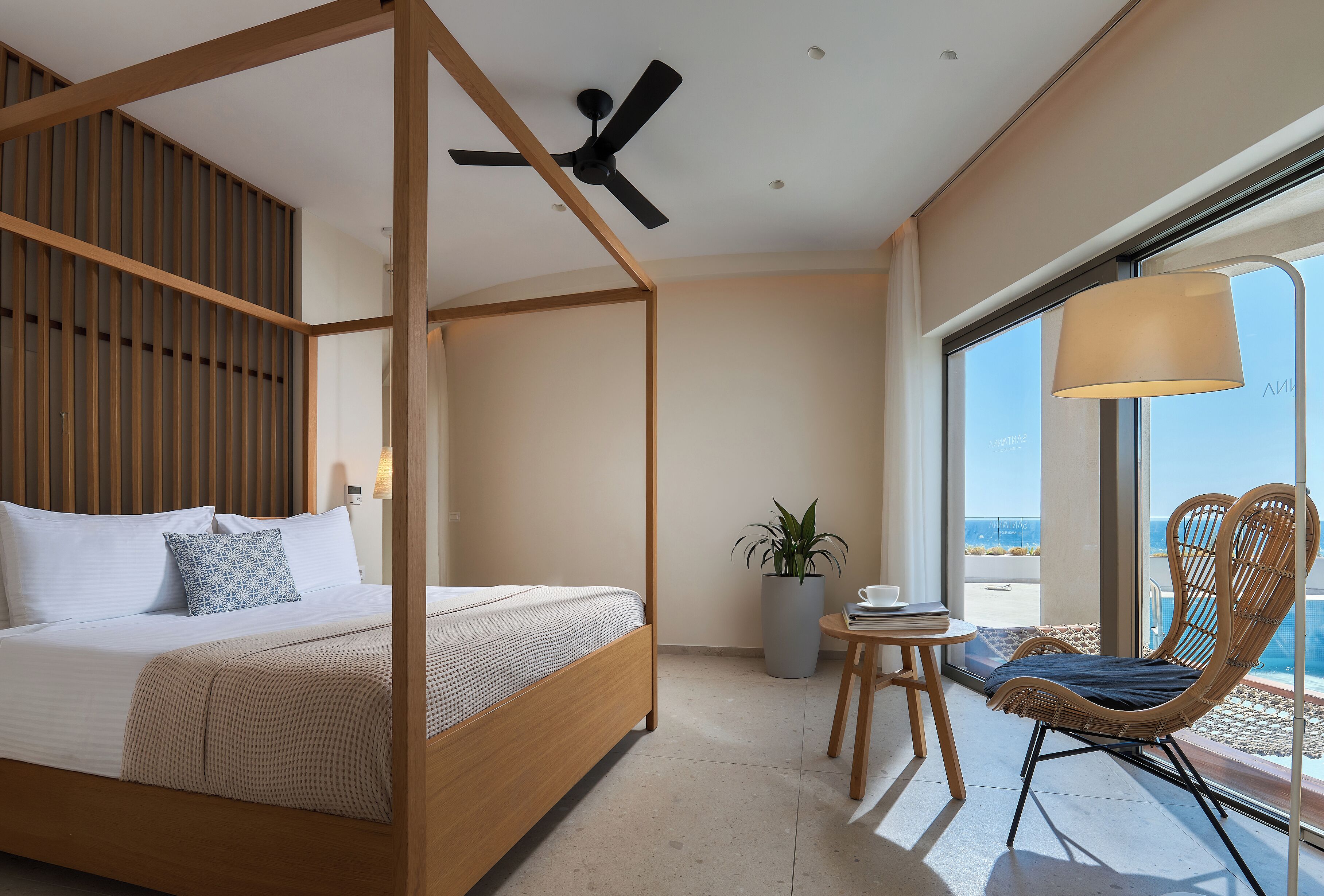 Una camera da letto con un letto a baldacchino, una ventola a soffitto, una pianta in vaso, una sedia in vimini e vista sull'oceano.
