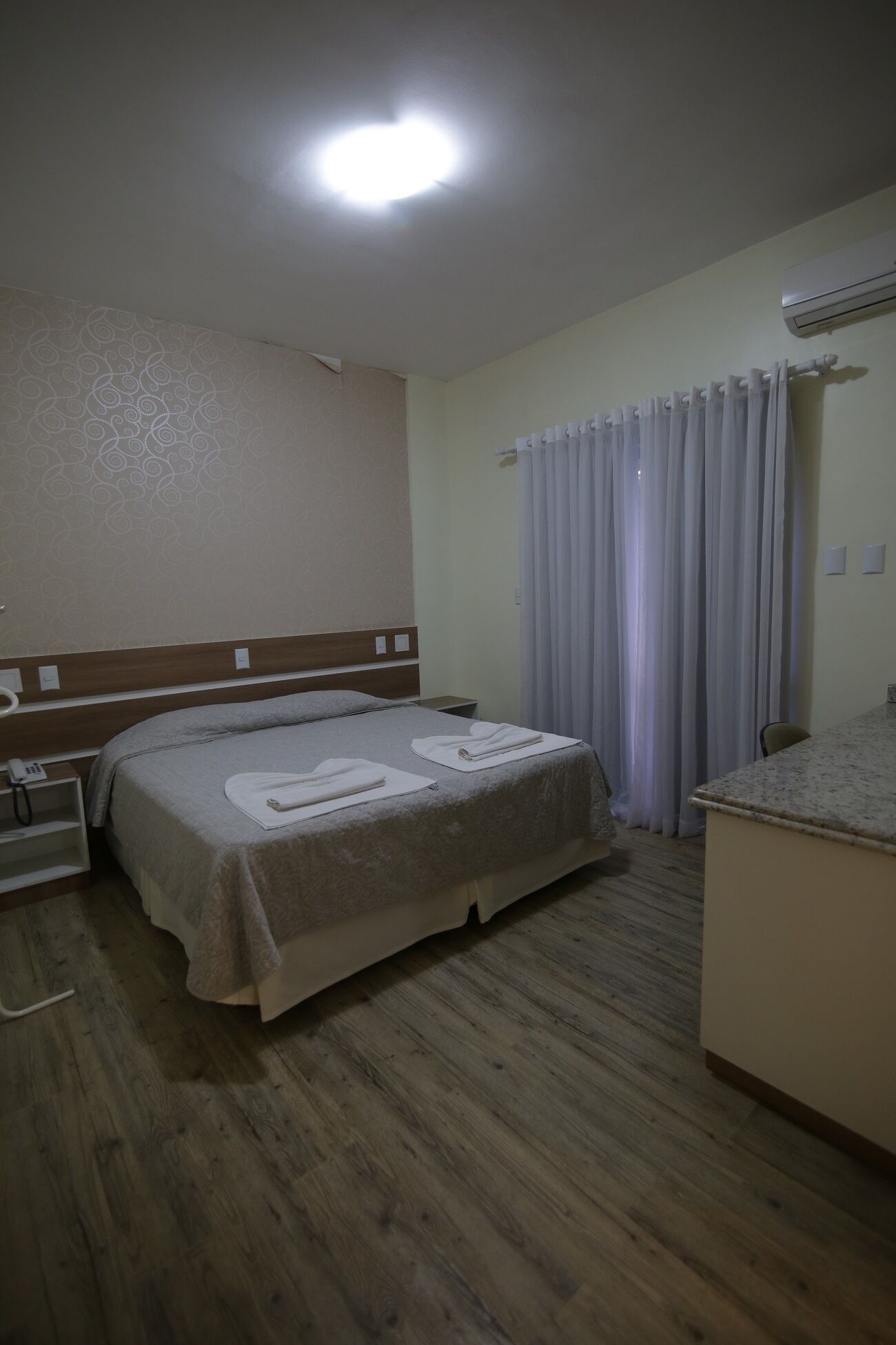 Ein Hotelzimmer mit einem Bett, einem kleinen Nachttisch, einem Schreibtisch mit Stuhl und einem Fenster mit Vorhängen.