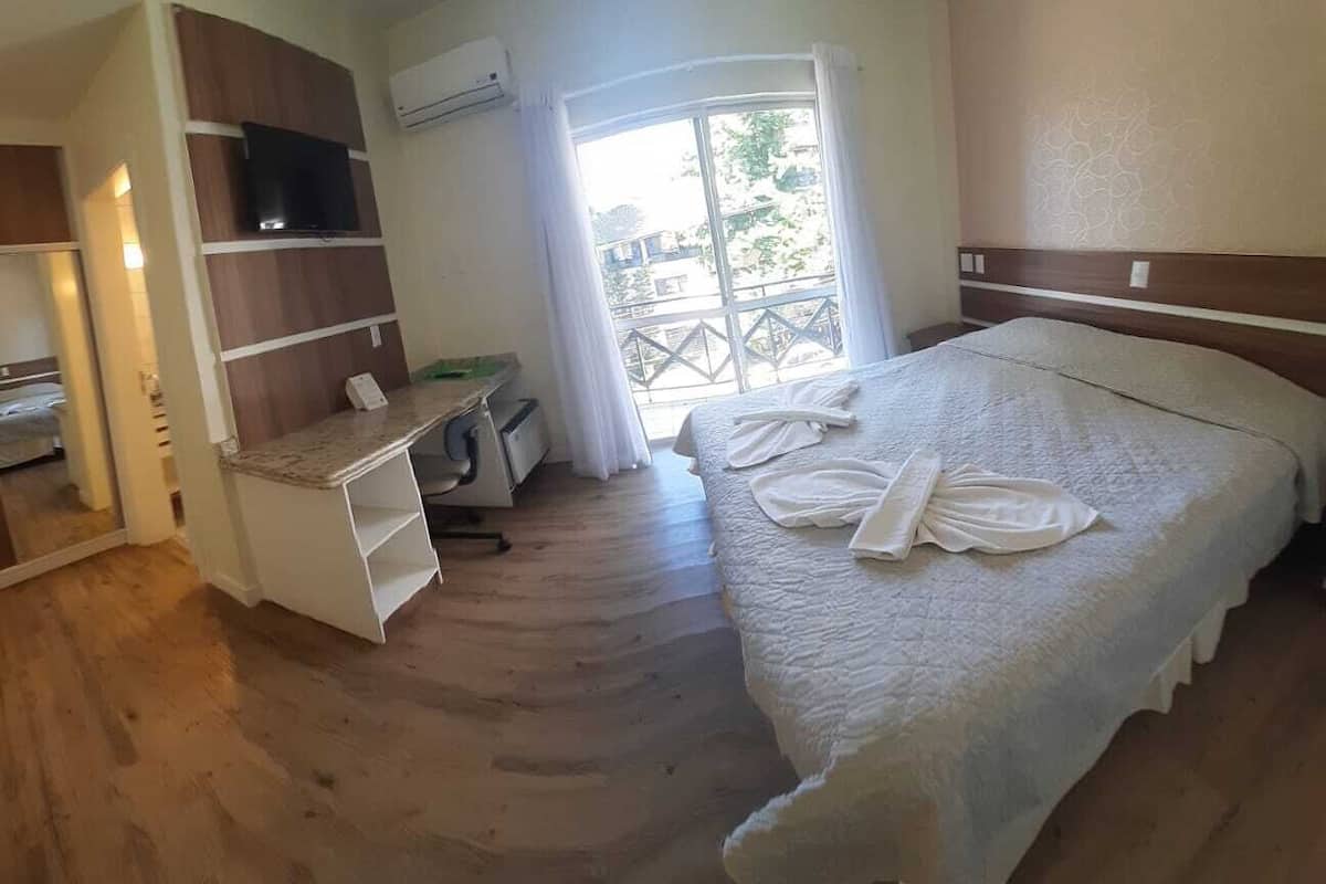 A bedroom with a bed, a desk, a chair, a TV, and a window with curtains.