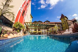 A swimming pool with a tiled deck, surrounded by buildings with a mix of architectural styles, including a red building with white trim and a yellow building with a red roof. - Hotel Kirst (Piratuba)