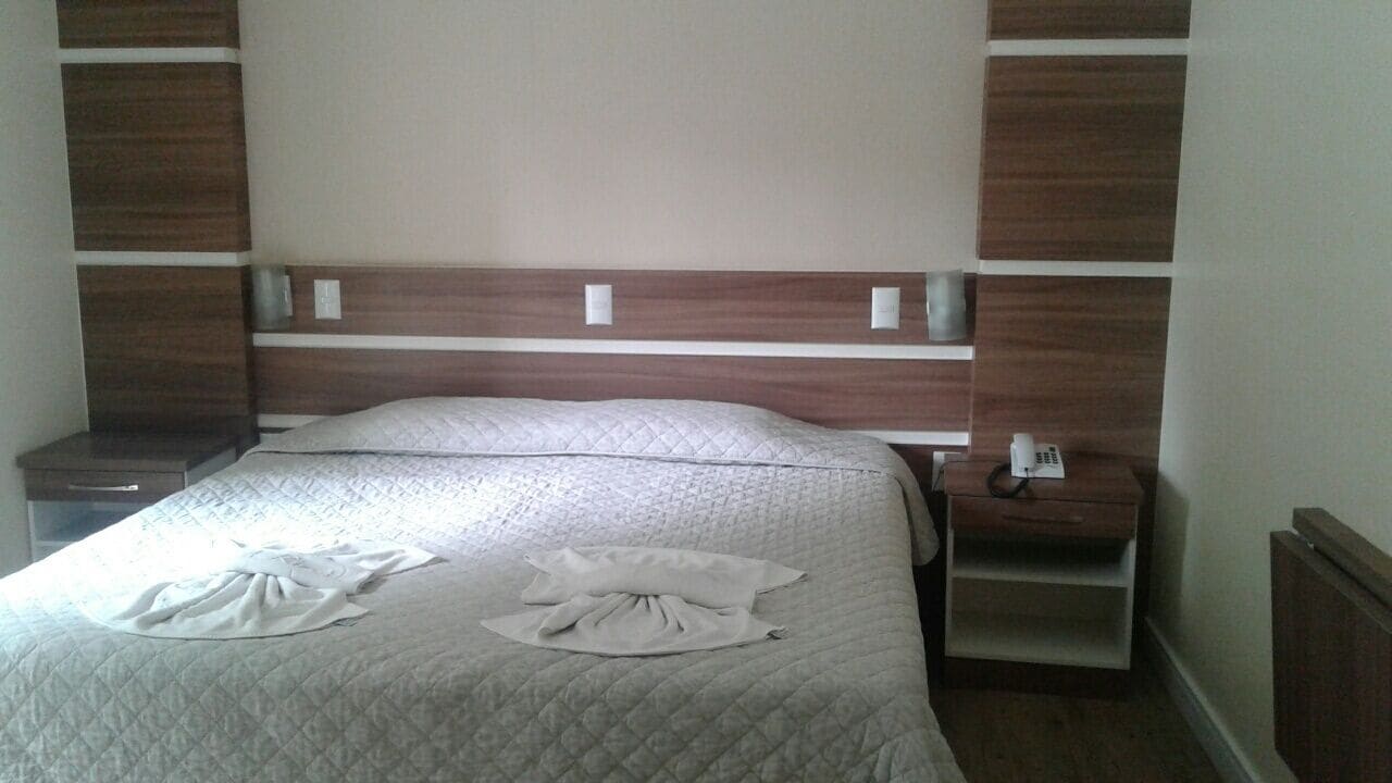 Una cama bien hecha con colcha acolchada y dos toallas blancas dobladas. Tiene un cabecero de madera con un panel de color claro y una mesita de noche con una lámpara y un teléfono.