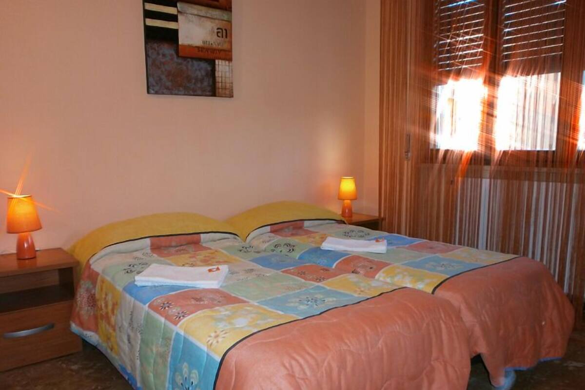 Une chambre à coucher avec un lit, deux lampes de chevet, une fenêtre avec des stores et une étagère fixée au mur, sur laquelle sont posés des livres.