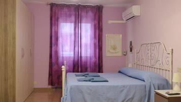 A bedroom with a bed, a nightstand, an air conditioning unit, and a window with purple curtains.