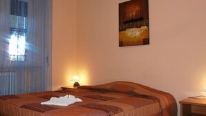 A hotel room with a bed, a nightstand, a lamp, a window with blinds, and a painting on the wall. - Airport Pisa Rooms (Pisa)