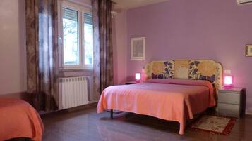 A bedroom with two beds, a radiator, a window with curtains, a chandelier, and a bedside table with a lamp.