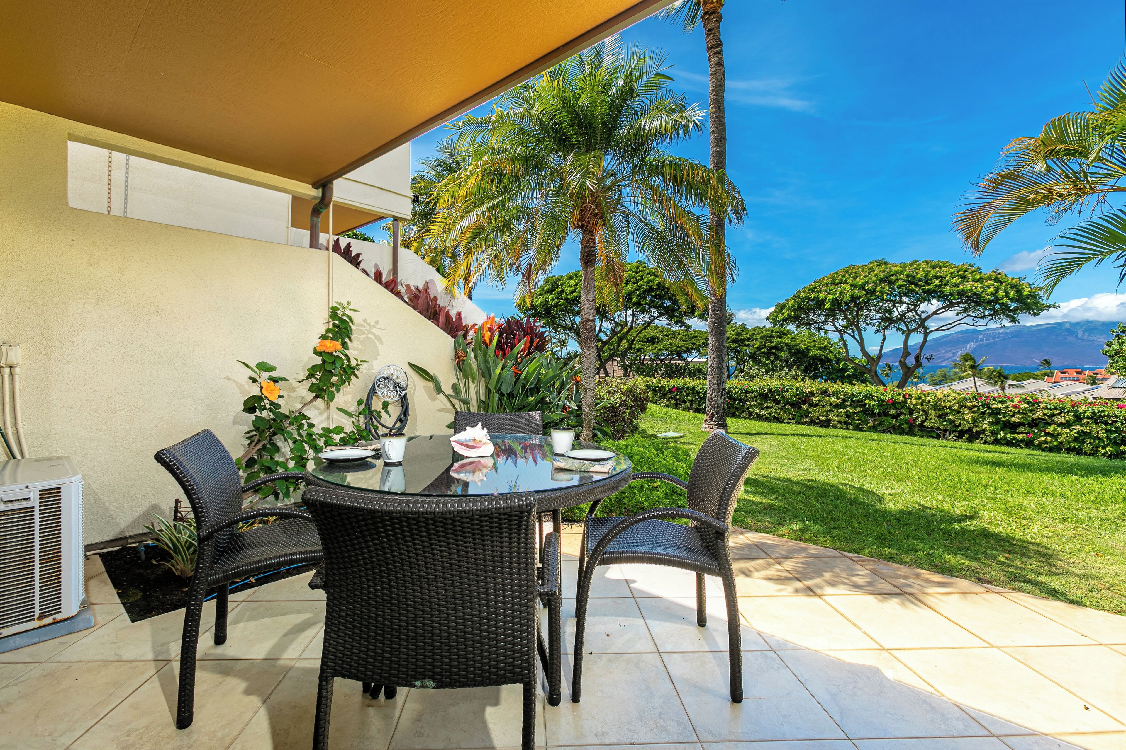 A patio with a round table and chairs, surrounded by potted plants and a view of a lush garden and mountains.