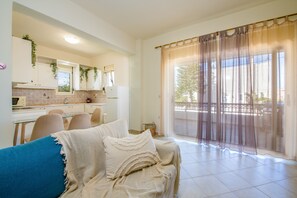 A modern living room with a sofa, a dining area with chairs, a kitchen with cabinets and a window, and a balcony with a view. - Dione Apartment with Terrace near Kalathas beach (Chania)