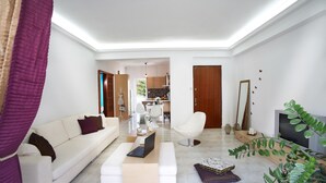 A modern living room with a white sofa, a coffee table with a laptop, a white armchair, and a TV. There is a kitchen visible in the background. - Dream City Apartment near Beach & Venetian Port (Chania)