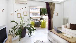 A modern living room with a sofa, a coffee table, a TV, and a balcony with yellow chairs and a table. - Dream City Apartment near Beach & Venetian Port (Chania)