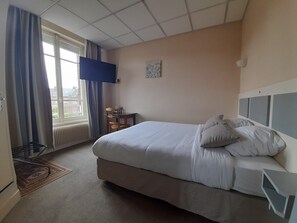 Une chambre d’hôtel comprenant un lit, un bureau, une chaise, un radiateur, une fenêtre avec des rideaux et un téléviseur fixé au mur.
