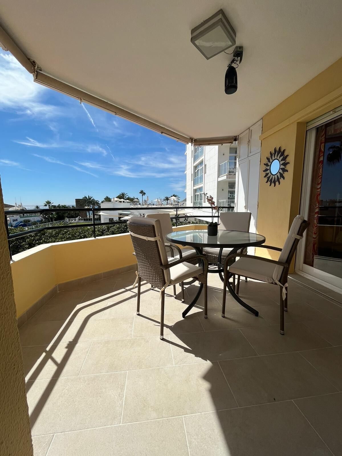 Un balcón con una mesa redonda y sillas, vista al mar, y un edificio con un adorno de sol.