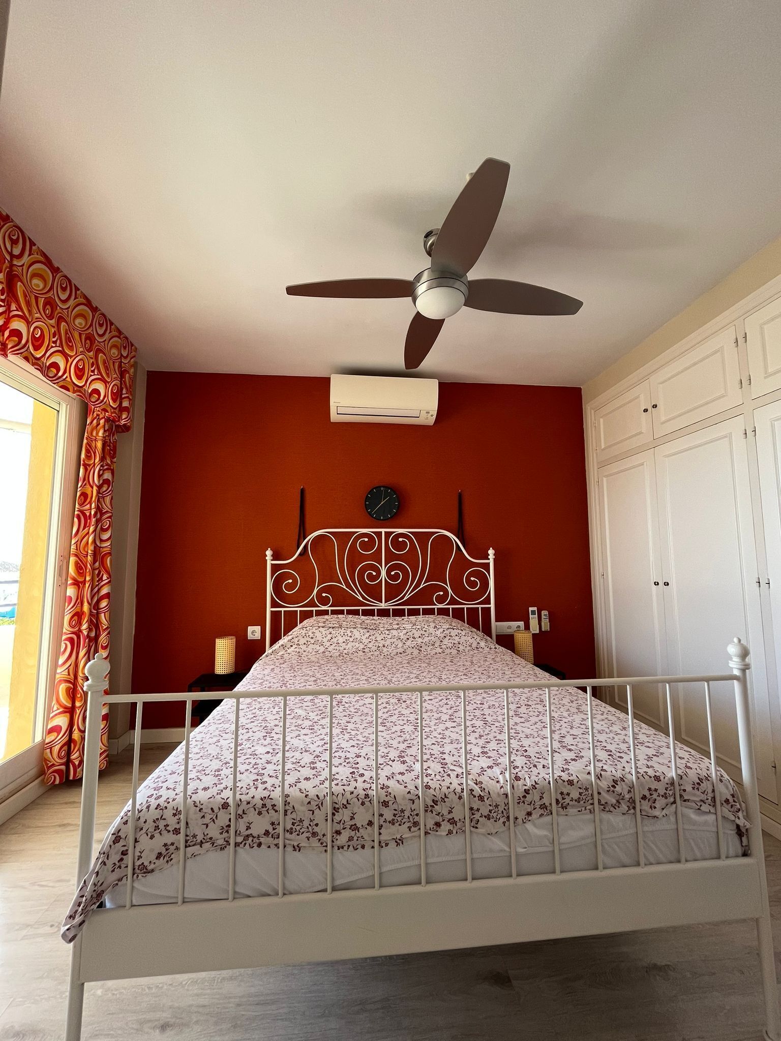 A bedroom with a bed, a ceiling fan, an air conditioner, a clock, and a wardrobe.