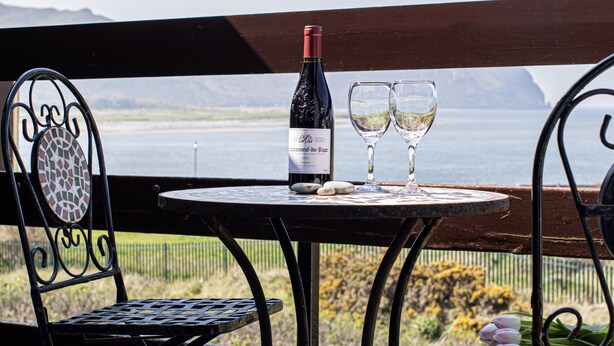 A table with a bottle of wine and two glasses, set on a balcony overlooking a body of water and mountains. - Fantastic Location with Stunning Estuary Views (Deganwy)