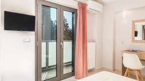 Une chambre moderne dotée d’une porte-fenêtre coulissante donnant sur un balcon, d’un téléviseur à écran plat fixé au mur, d’un bureau blanc avec une chaise et d’une grande fenêtre offrant une vue sur les arbres.