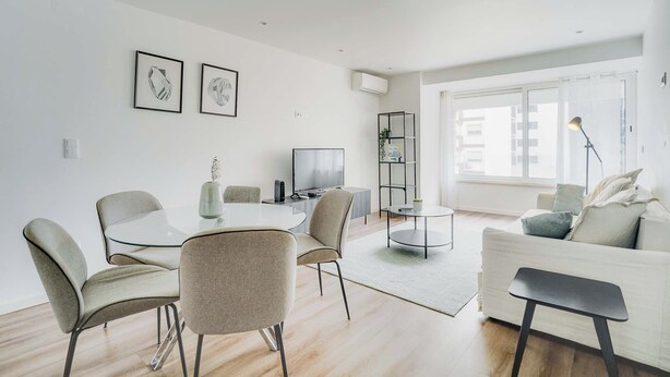 A modern living room with a glass dining table, comfortable seating, a flat-screen TV, and a large window providing natural light. - Savory Blue Apartment, Wifi, Garage, Lift!New! (Lisboa)