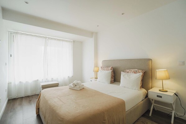 A hotel room with a large bed, two bedside tables with lamps, a bench, and a window with sheer curtains.