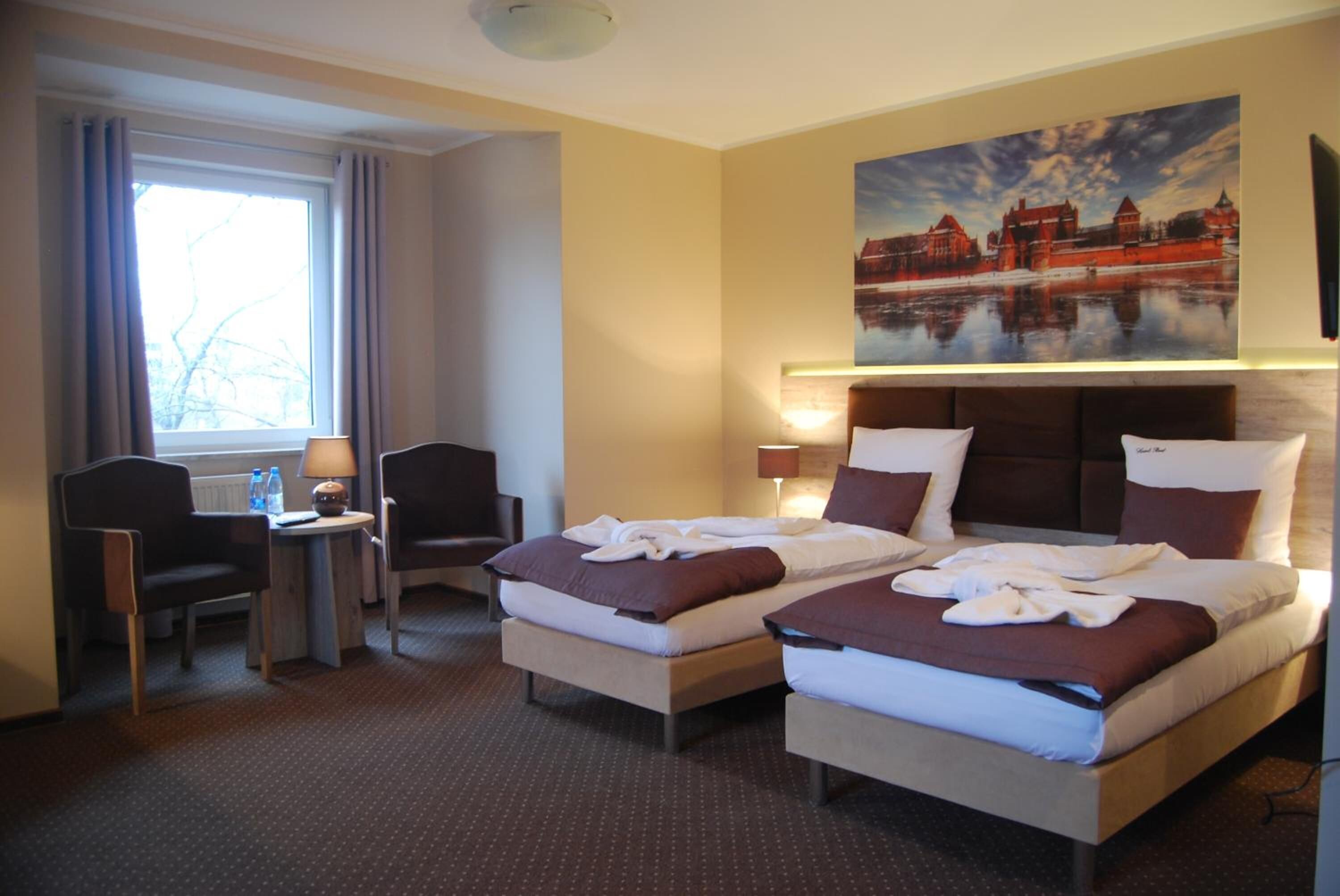 A hotel room with two beds, a desk, chairs, a window with curtains, and a painting of a castle on the wall.