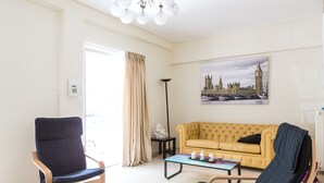 Ein Wohnzimmer mit einem gelben Sofa, zwei schwarzen Sesseln, einem Glastisch, einer Stehlampe und einem Stadtbild.