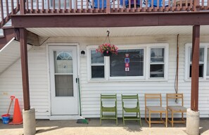 A porch with a wooden railing, a white door, and a window with blinds. There are three chairs and a broom on the porch.