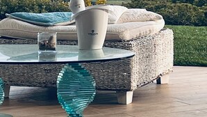 Een moderne buitenzitplaats met een rieten bank, een glazen bijzettafeltje en decoratieve glazen voetstukken.