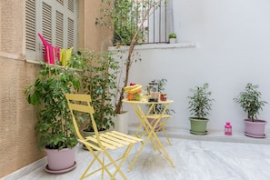 A small outdoor seating area with a table and chairs, surrounded by potted plants and a window with shutters. - Grandma's Chic & Bright Athenian Home with a Patio (Athens)