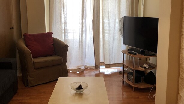 Una sala de estar con un sofá, una mesa de centro, un televisor sobre un mueble bajo y una ventana con cortinas.