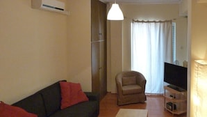 Una sala de estar con un sofá oscuro, una mesa de centro clara, un sillón color beige, un televisor sobre un mueble bajo y una lámpara.