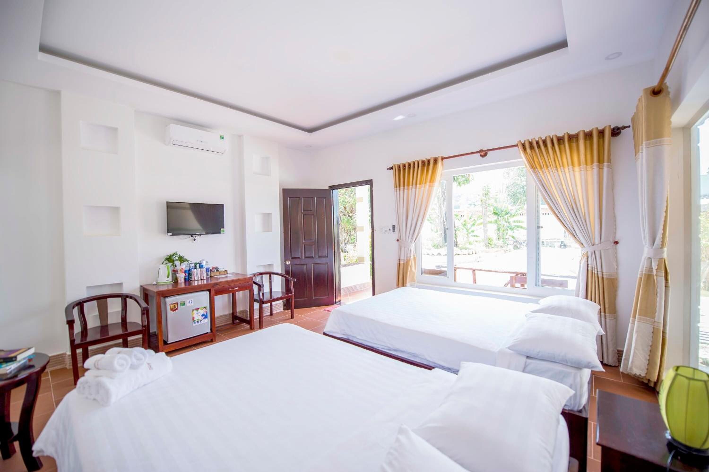 A hotel room with two beds, a desk with a television, a minibar, and a door leading to an outdoor area.