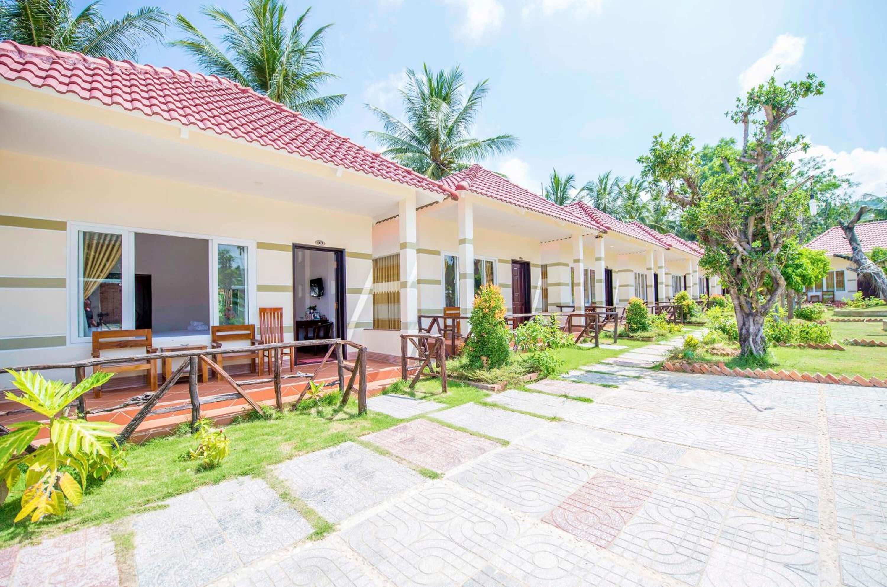 A row of hotel rooms with red-tiled roofs, wooden furniture, and a paved courtyard with palm trees.