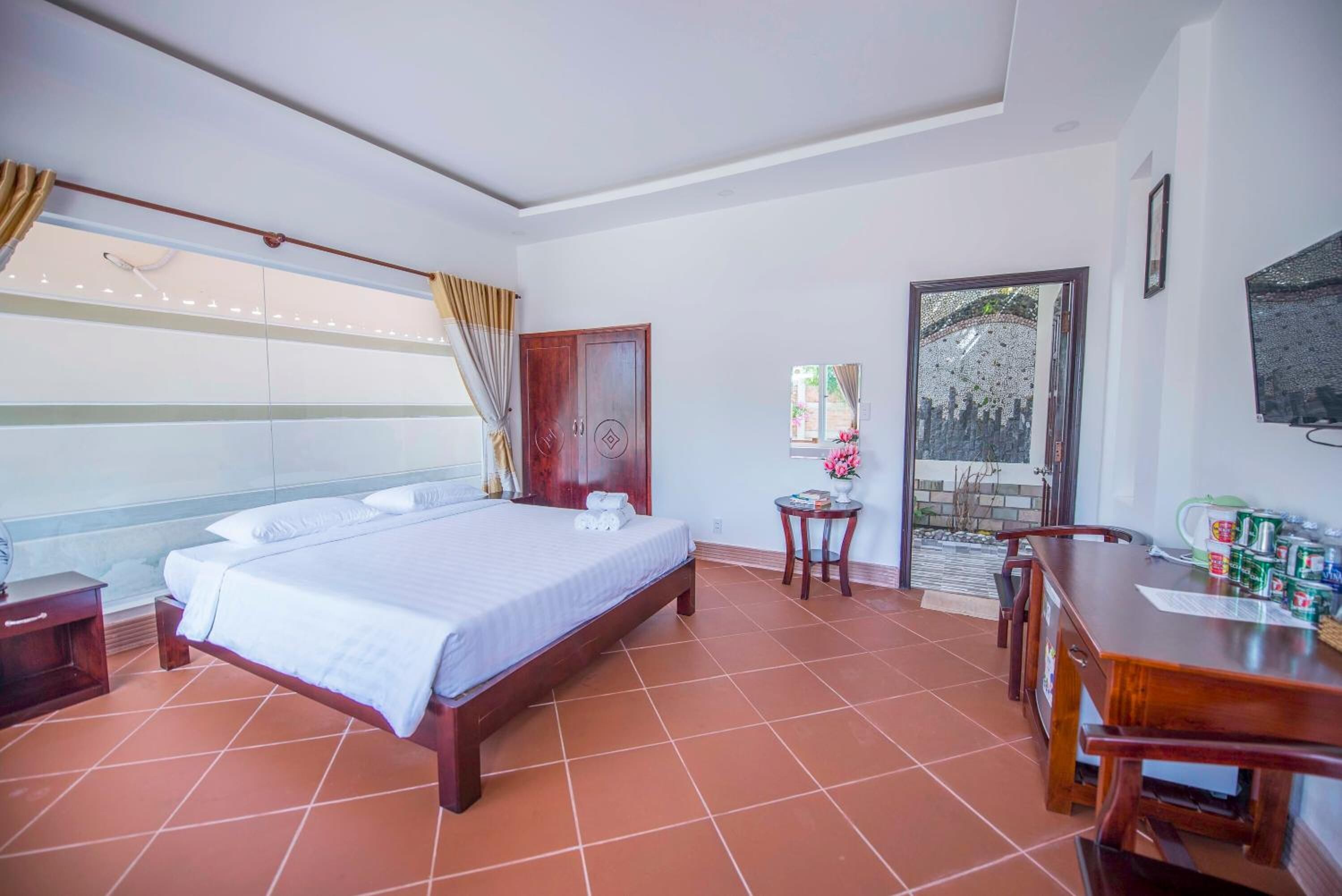 A hotel room with a large bed, a wooden desk, a small table, and a door leading outside.