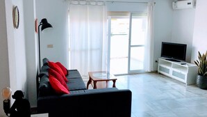 Een moderne woonkamer met een bank, een televisie op een witte kast en een glazen schuifdeur naar een buitenruimte.