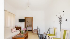 A cozy living room with a sofa, coffee table, and chairs. A TV is mounted on the wall, and a door leads to a bedroom.