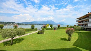 A well-maintained garden with a pathway, a building with balconies overlooking a lake, and mountains in the background. - Sogno del Lago: in front lane of the beach in Manerba del garda (Manerba del Garda)