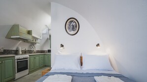 Une chambre à coucher comprenant un lit, deux tables de chevet, une commode, un tableau accroché au mur, et un coin cuisine avec des meubles verts et une cuisinière.