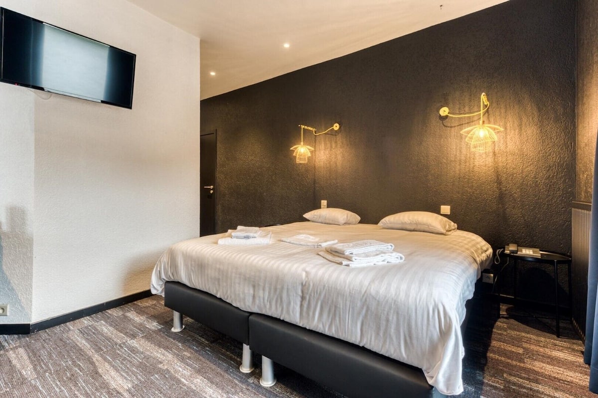A modern hotel room with a large bed, two bedside tables, a flat-screen TV mounted on the wall, and a unique wall-mounted light fixture.