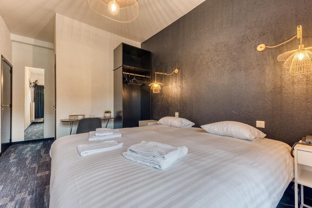 A hotel room with a large bed, a desk, a chair, and a wall-mounted light fixture.