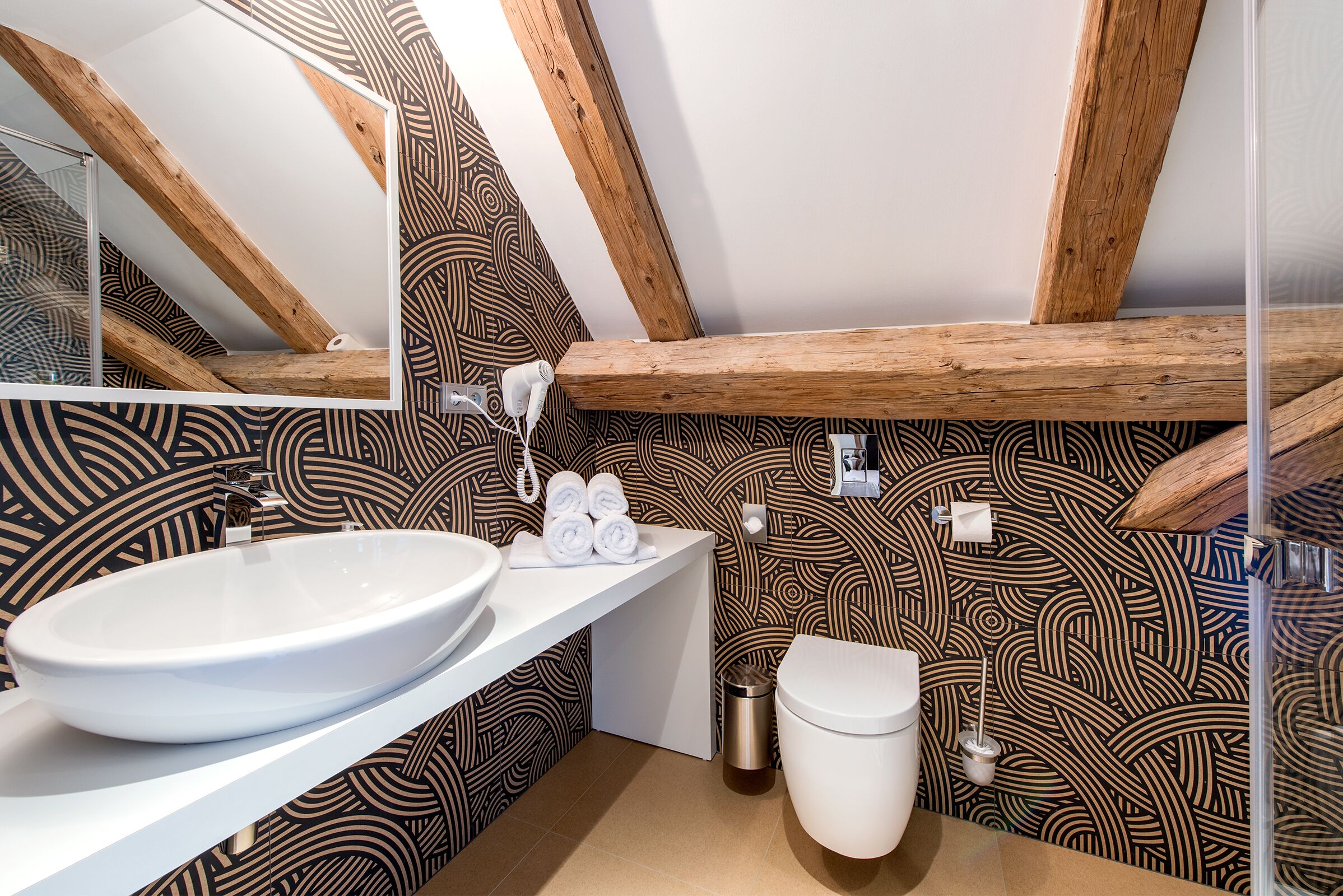 Une salle de bain avec un lavabo blanc, des toilettes et un miroir. Les murs sont recouverts d’un papier peint à motifs, et le plafond est orné de poutres en bois.