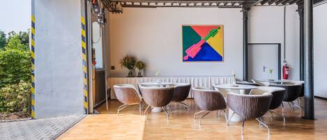 Un área de estar interior con sillas de mimbre y una mesa, un cuadro abstracto y colorido en la pared, y un extintor.