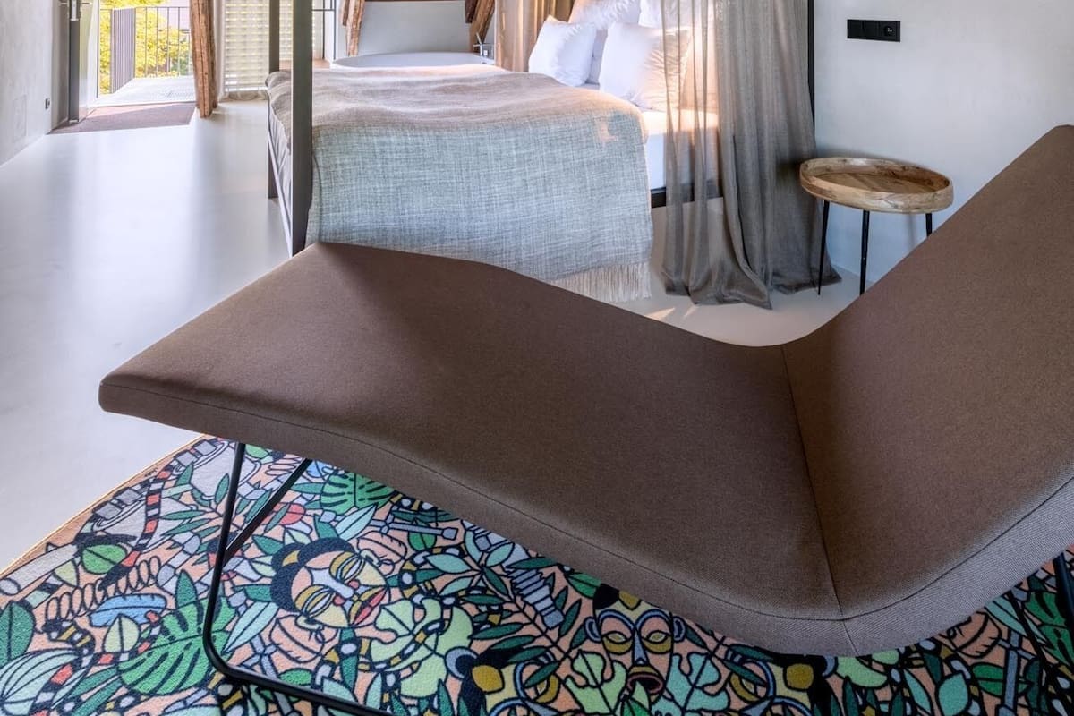 A bedroom with a bed, a bench, a hanging light fixture, and a view of the outdoors.