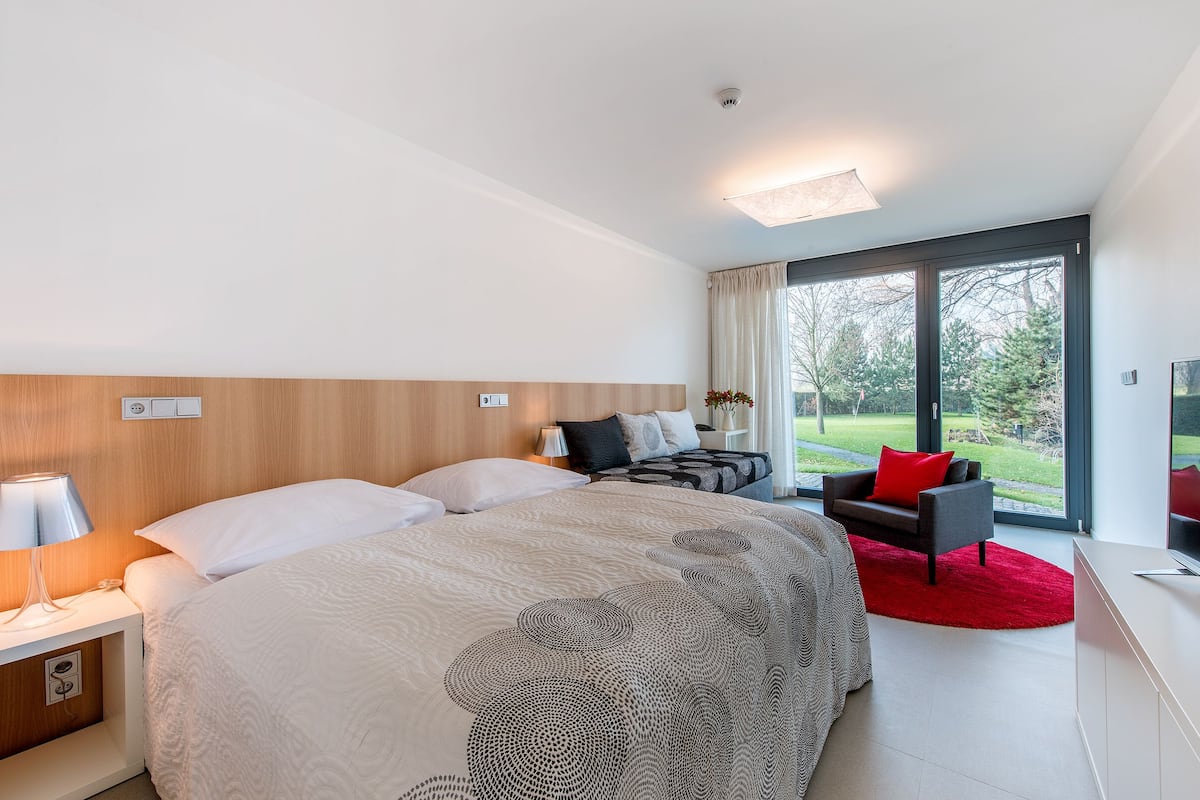 A modern hotel room with a large bed, a desk, a chair, and a view of the outdoors.