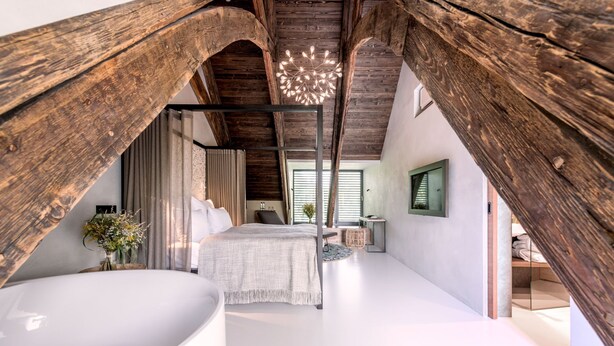 Une chambre avec une grande baignoire blanche, un lit recouvert de linge de maison blanc et une structure de plafond en bois.