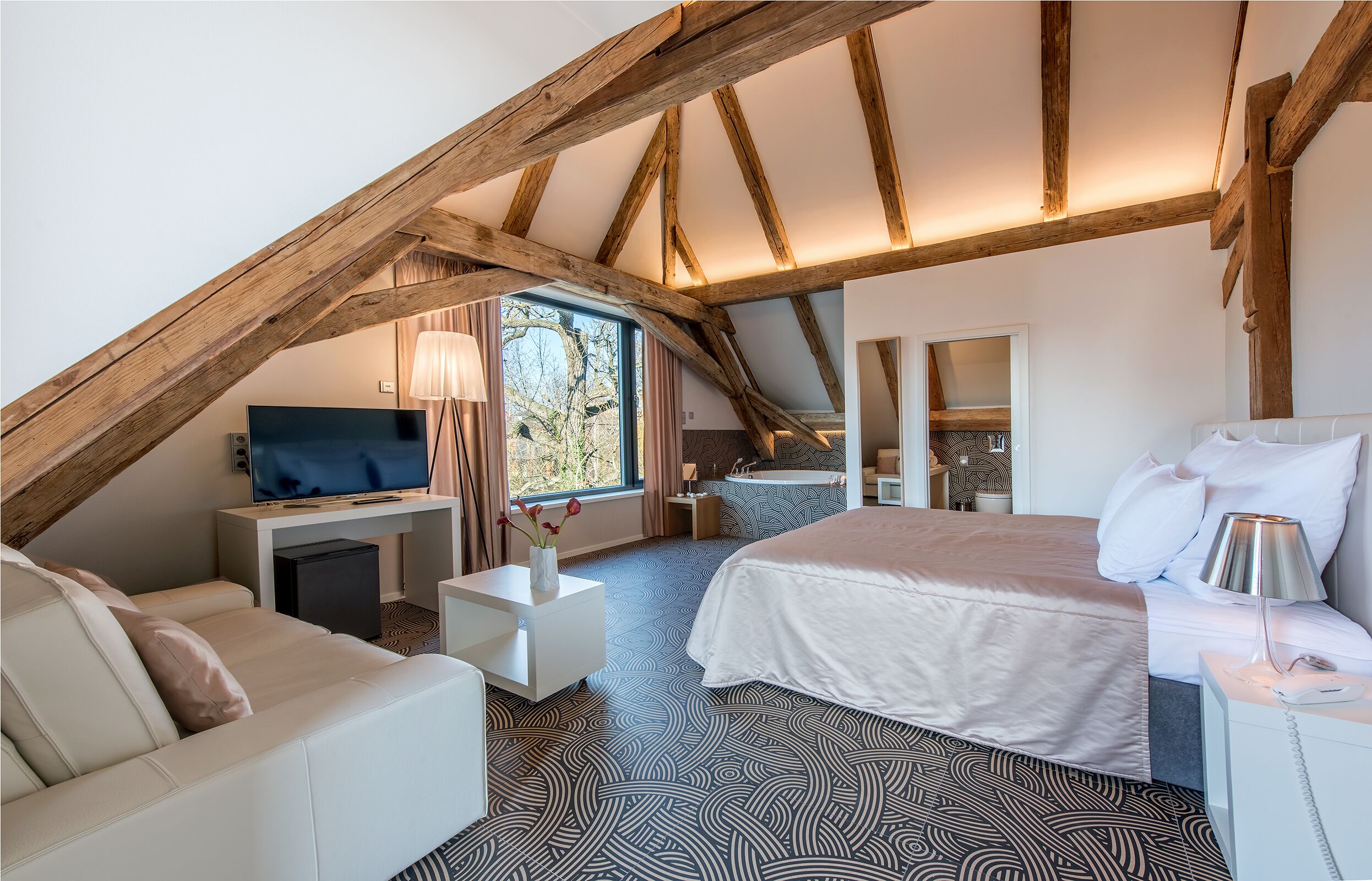 A bedroom with a sloped ceiling, a bed, a desk, a TV, and a window with curtains.