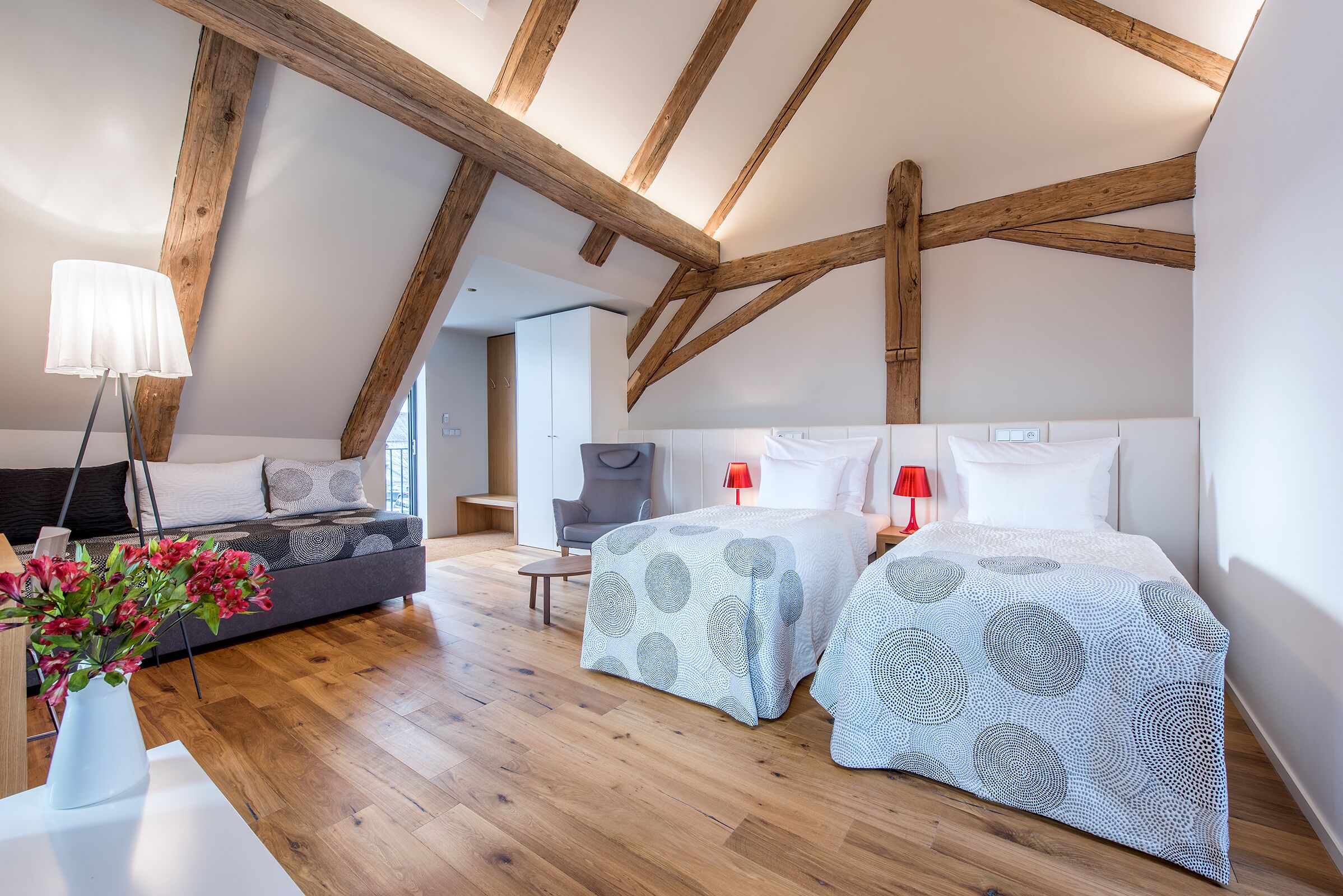 Un dormitorio moderno con vigas de madera a la vista, un sofá, dos camas con ropa de cama estampada, un jarrón de flores y una lámpara roja.