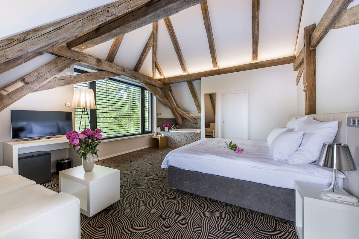 A bedroom with a sloped ceiling, a bed, a desk, a TV, and a window with blinds.