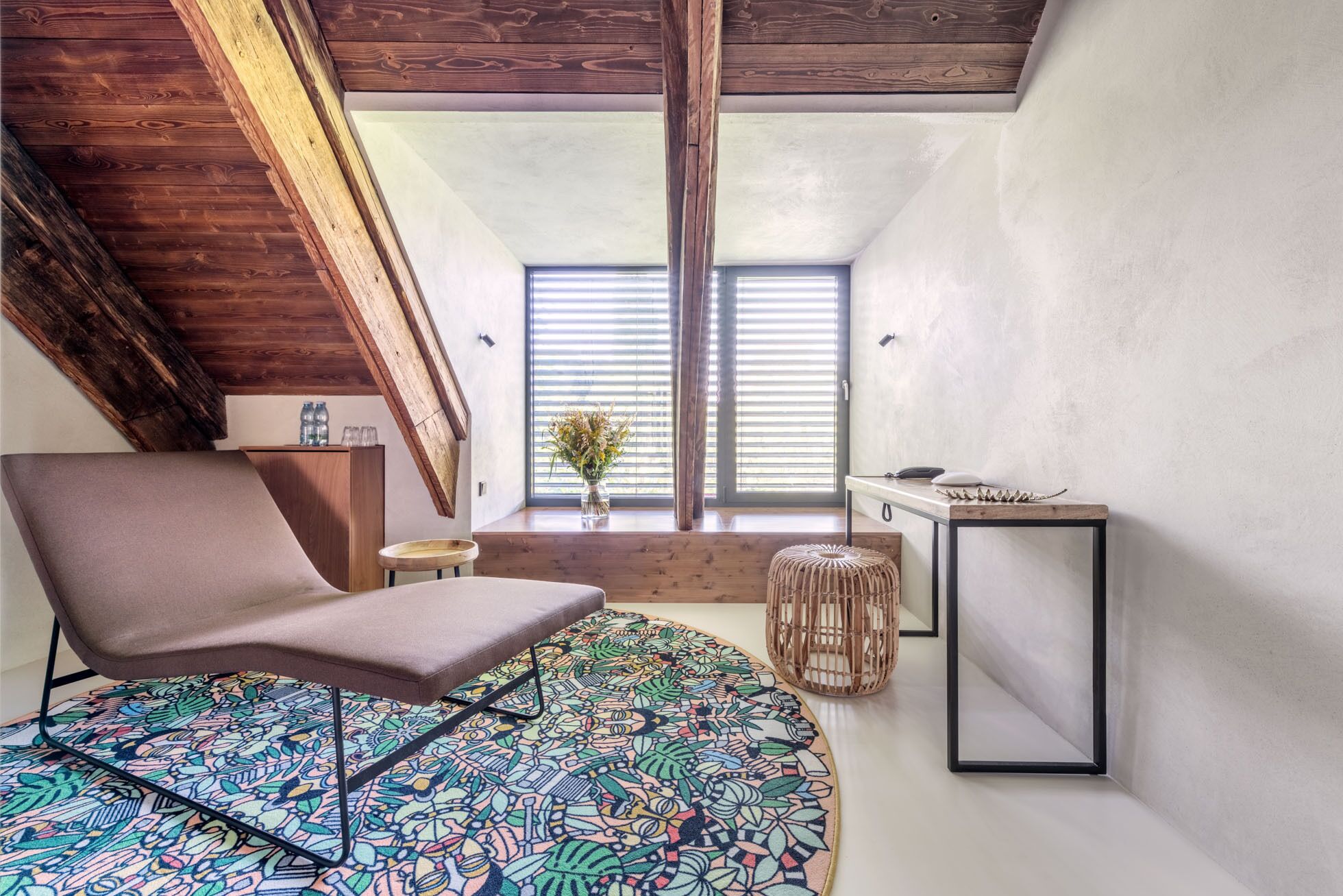 A loft-style bedroom with a sloped ceiling, a large window with blinds, a bed with a colorful patterned rug, a chair, a small table, and a bedside table with a lamp.