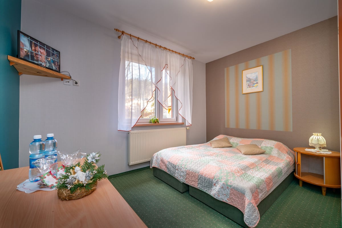 Een slaapkamer met een bed, een raam met gordijnen, een radiator, een nachtkastje met een lamp en een wandplank met een foto.